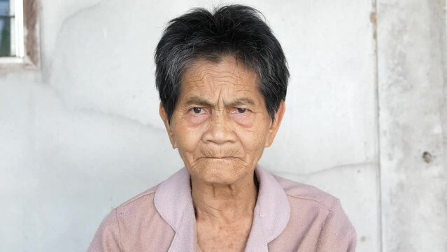 Asian elderly native sad woman looking to camera with serious face isolated concrete grey wall background. Concept of people rural lifestyle, unhappy expression.