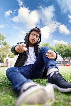 Wide angle shot of asian teenager sitting in lawn green grass pointing finger and look at camera in the park blue sky background