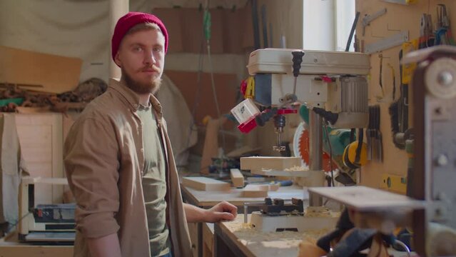 Young bearded carpenter in red beanie hat blowing sawdust from wooden plank and posing for camera in woodworking workshop