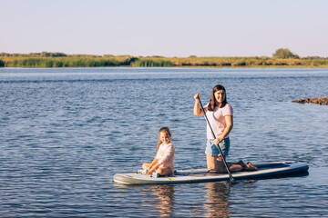 Fototapeta na wymiar Young daughter and mother sitting together on sup board rowing with paddle on little lake with green reeds and trees in background. Active holidays. Inculcation of love for sports from childhood.