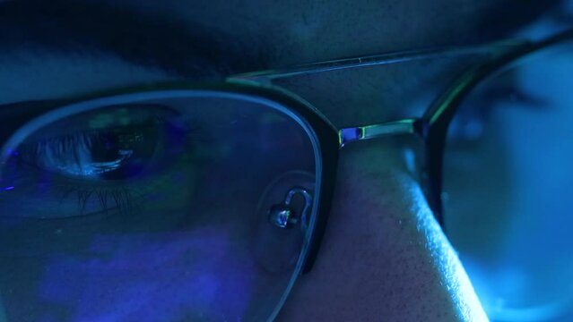 Blue light of a computer screen reflected on a young male glasses. Extreme close up view, macro shot in 4k of young male looking at screen monitor.
