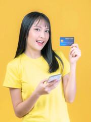 beautiful Asian young woman cute girl with bangs hair style in yellow shirt holding credit card and smartphone ,mobile phone uses a online banking for pay online shopping isolated on yellow background