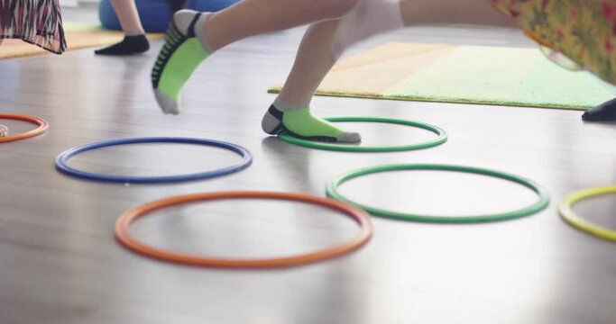 Small nursery school children with female teacher on floor indoors in classroom, doing exercise. Jumping over hula hoop circles track on the floor.
