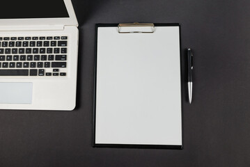 Top view photo of black clipboard folder with white paper sheet, pen and laptop on isolated black background