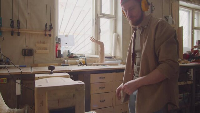 Woodworker in safety earmuffs using thickness planer while working with plank in carpentry workshop