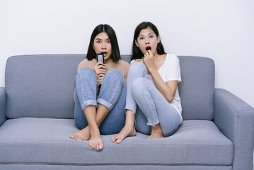 Scared Young Asian Lesbian couple watching horror movie on tv holding remote control sitting on couch at home. Girls feeling fear or surprise during thrilling scary film moment, emotional reactions.
