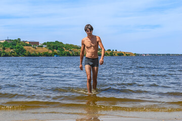 A young slender man in swimming trunks and sunglasses walks out of the river on Strilka beach in...