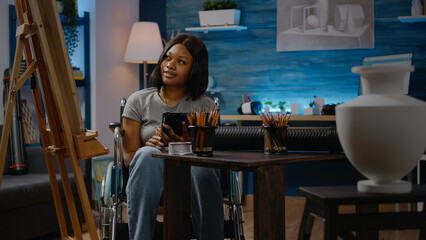 Obraz premium Young black artist in wheelchair using digital tablet sitting in art studio. African american adult creating masterpiece of vase design to draw with pencil on white canvas and easel.