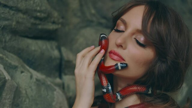Portrait close-up Gothic fantasy woman dark queen of snakes and poisons. Girl princess holds in hands, plays with aspred snake sinaloa. Lady vampire, black dress. Royal milk snake crawls on skin face.