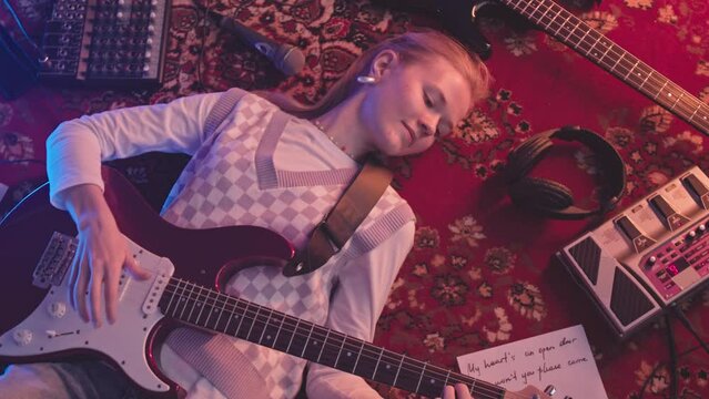 From above medium slowmo of young y2k woman smiling while playing electric guitar lying on retro red carpet with love song lyrics all over place