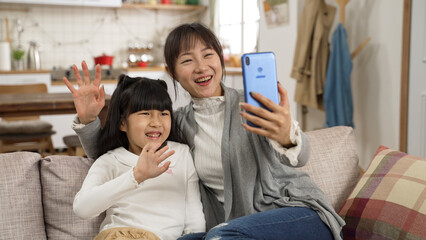 happy asian mom and girl talking to friends through video conferencing at home. the woman holds mobile phone and sits close to her daughter on sofa