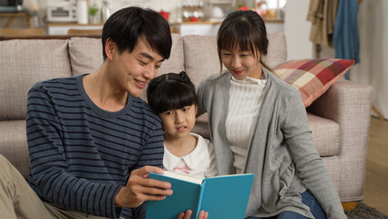 cheerful asian family of three enjoying reading together in the living room at home. the smiling...