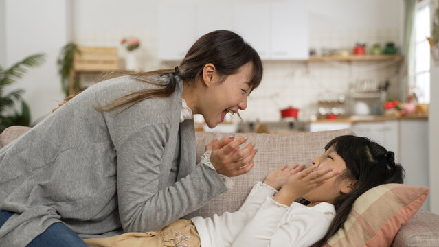 loving Asian mother touching her daughter’s face while she is lying on couch making funny silly face to her in the living room at home