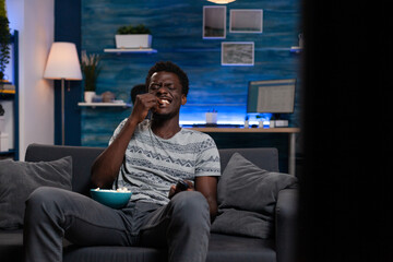 Smiling young person resting on sofa holding bowl with popcorn snacks while watching comedy movie...