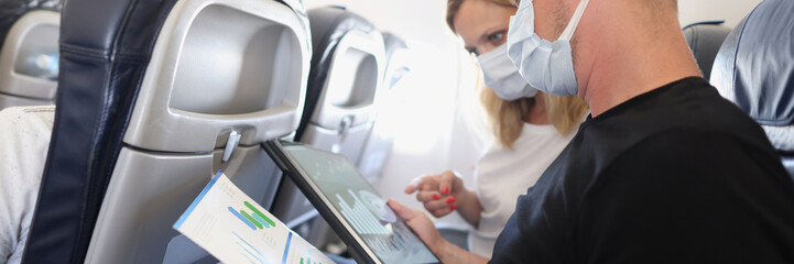 Man and woman in protective medical masks flying in airplane and discussing diagram on documents