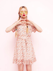 Young beautiful smiling female in trendy summer red dress. Sexy carefree blond  woman posing near wall in studio. Positive model having fun. Cheerful and happy in sunglasses. Makes heart sign