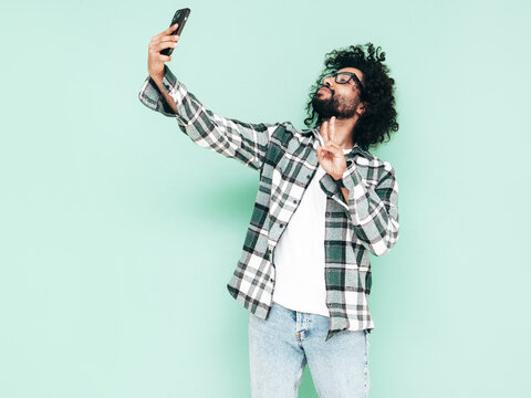 Portrait of handsome smiling hipster  model.Unshaven Arabian man dressed in summer shirt and jeans clothes. Fashion male with long curly hairstyle posing near blue wall in studio. Taking selfie photos