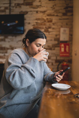 Chica joven guapa tomando té  en una cafetería muy acogedora con sudadera gris y mirando el smartphone