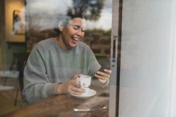 Chica joven guapa tomando té  en una cafetería muy acogedora con sudadera gris y mirando el smartphone