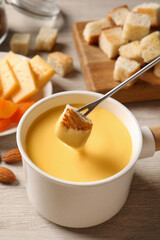Dipping piece of bread into tasty cheese fondue on light wooden table