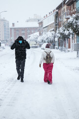 a man in black with mask and a woman in pink and white with bag walking over snow way with trees, buildings and cars