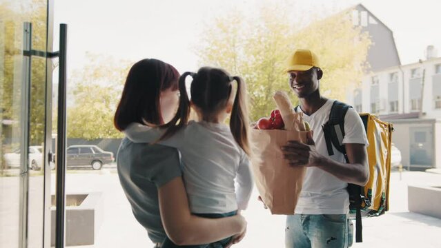 An African-American courier brings a package of food, fruits and vegetables to a beautiful woman with a child, a client in front of the house. Concept of express food delivery and online shopping.