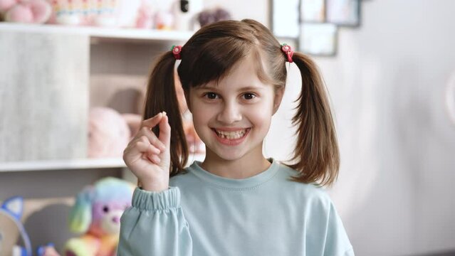 Portrait of cute little child girl is rejoices and showing her lost milk tooth and smiling to camera of toothless mouth while she standing in bedroom. Happy girl holding her fallen tooth in hand.