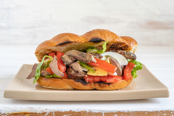 Challah sandwich with roasted liver, fresh tomatoes, boiled eggs on beige plate over wooden background.