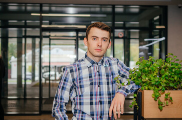 a guy in a shirt leans on a flowerpot and looks at the camera against the background of glass doors