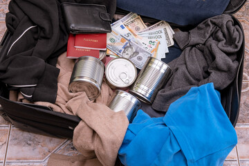 Clothes, canned food, money and documents are in a suitcase. Concept - collection of things during a natural disaster, emergency, quick evacuation during a disaster. - obrazy, fototapety, plakaty