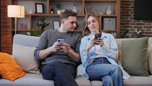 Couple sitting on couch together and using their phones, young couple lifestyle, leisure activities. The girl shows photos from social networks to her boyfriend. Dependence on mobile phones. 