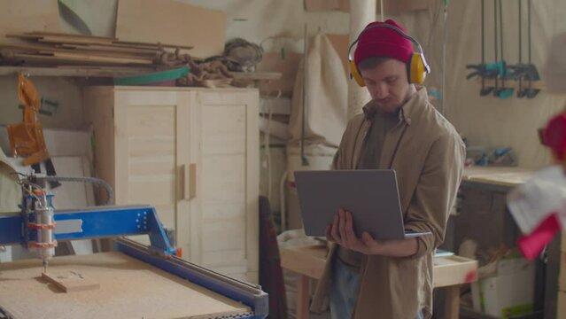 Woodworker in safety earmuffs standing in workshop and operating CNC machine with laptop