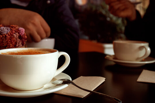 Close-up shot of cup of turkish traditional winter drink salep with cinnamon with male hand on blurred background. White ceramic mugs on plates and little spoon on table. Selective focus.