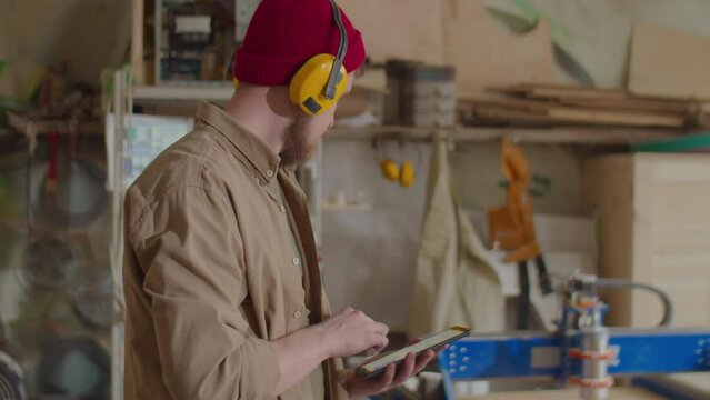 Woodworker in safety earmuffs using smartphone in workshop while operating woodworking CNC machine with mobile app