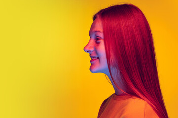 Side view portrait of red haired beautiful teen girl posing, smiling isolated over yellow studio background in neon light