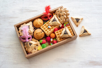 Sweet box with Hamantaschen cookies, candies, cupcakes, waffles for Jewish holiday Purim on white wooden background.