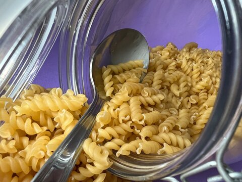 A tablespoon is in a glass jar with dry pasta. Lilac background.