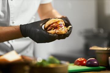 Crop view of head chef's hands in gloves holding tasteful cheeseburger with vegetables. Close up...