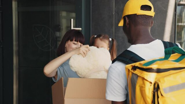 Courier bring a parcel to a little girl with mom at the door of the house. Opening the parcel, a happy child takes out of the box a fluffy teddy bear toy, rejoices in the surprise, family delivery.