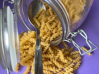 A tablespoon is in a glass jar with dry pasta. Lilac background.