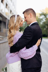 Perfect smiling couple of young bride with wavy fair hair in bridal dress, lilac cardigan standing hugging bridegroom.