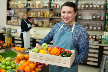 Smiling male salesman holds a wooden box with vegetables and fruits in the store.