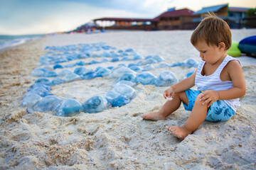 A boy is played with seashells and sand while sitting on a beach strewn with jellyfish near the sea © YouraPechkin