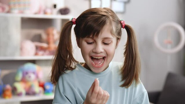 The child's milk tooth fell out. The pleased girl pulls out a milk tooth from his mouth and is surprised. Cute little child girl showing her lost milk tooth and smiling to camera standing in bedroom
