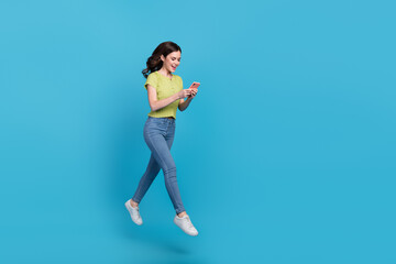 Full length photo of cute young brunette lady run look telephone wear t-shirt jeans sneakers isolated on blue background