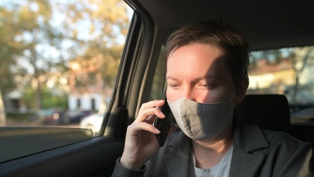 Serious businesswoman with protective face mask talking on mobile phone from the backseat of the car