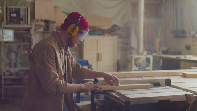 Woodworker in safety earmuffs and glasses using power jointer machine while working with board in carpentry workshop