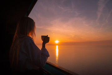 Happy young woman in a white shirt is drinking coffee from a white cup on the balcony, watching the...