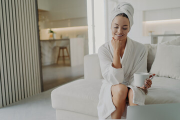 Happy woman in white bathrobe drinking tea and relaxing on sofa after taking shower or bath - obrazy, fototapety, plakaty