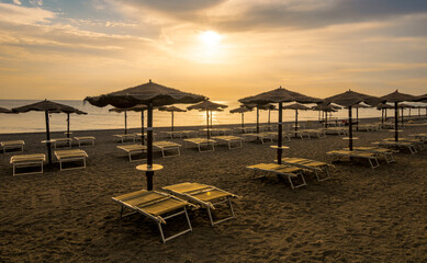  empty beach during beautiful sunrise or sunset with chaise loungues and nice umbrellas with blue sea, sun glow and amazing cloudy sky on thr background